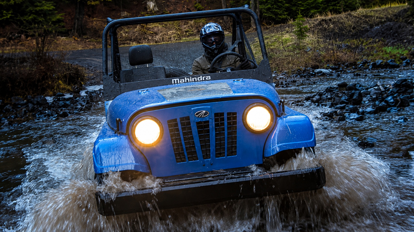 A blue Mahindra off road vehicle driving through a shallow, muddy stream