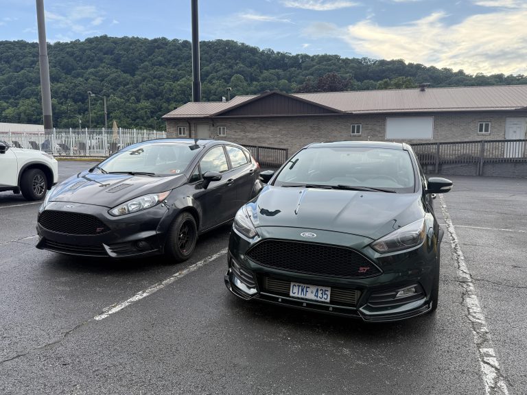Two dark hatchback cars parked side-by-side in an outdoor parking lot with a building and hill backdrop