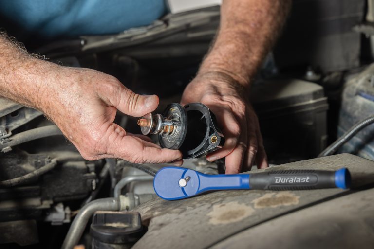 Mechanic holding a car thermostat in an engine bay next to a blue ratchet