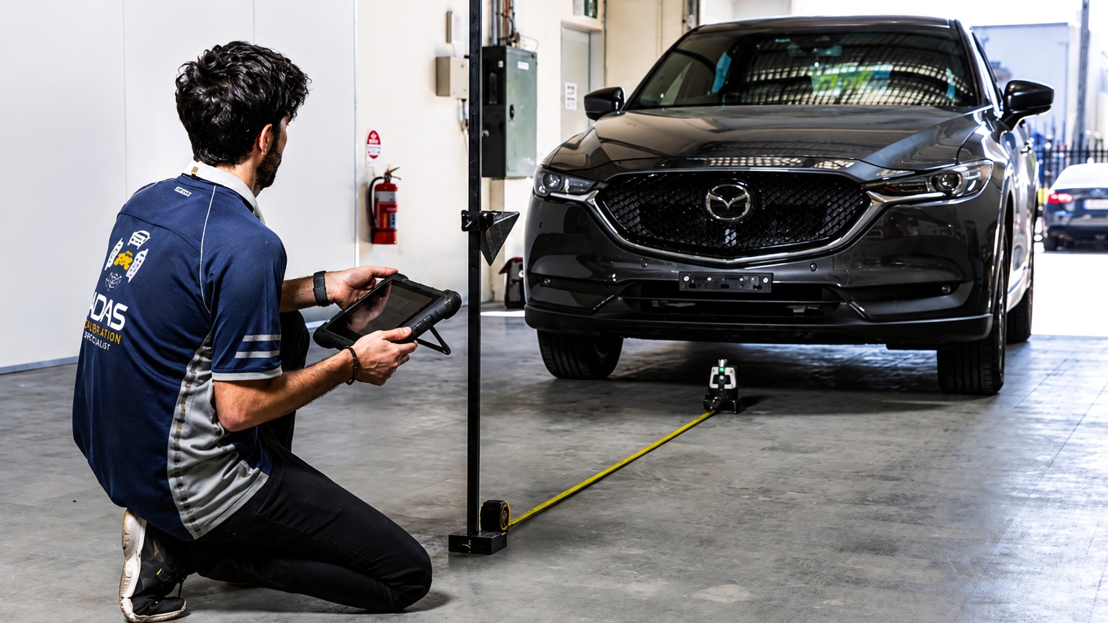 technician kneels in a garage, using a tablet to calibrate a dark SUV's ADAS sensors