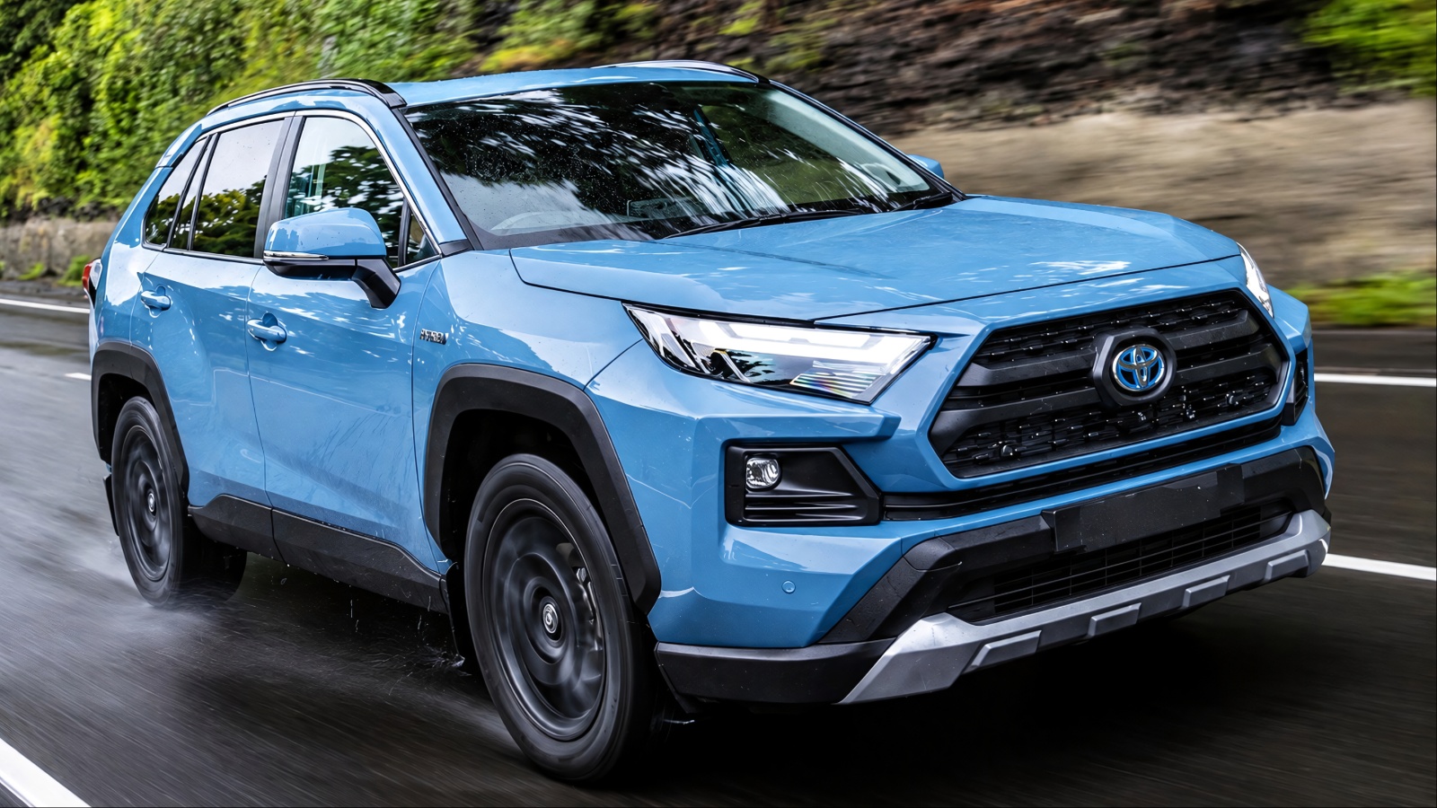 blue Toyota RAV4 SUV driving on a wet road next to a rocky cliffside