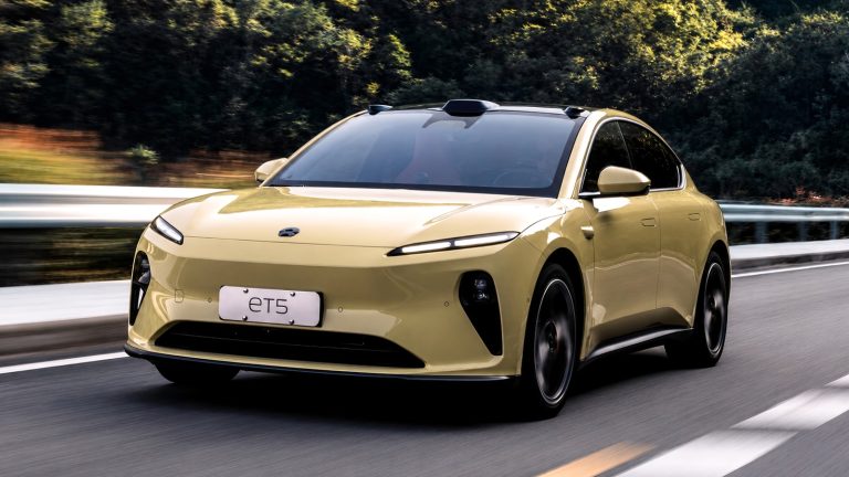 yellow electric sedan driving on a paved road surrounded by green trees