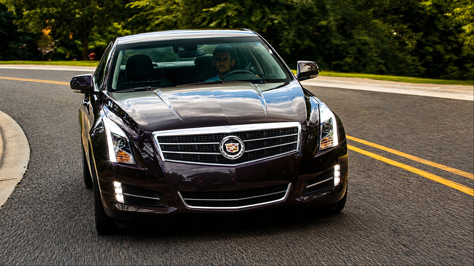 dark purple Cadillac sedan driving on a winding paved road surrounded by green trees