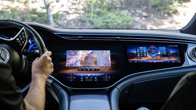 A driver's hands on a steering wheel facing a wide digital dashboard with multiple screens