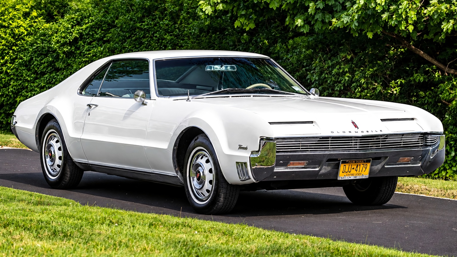 A sleek white vintage coupe parked on a paved driveway beside green grass and trees
