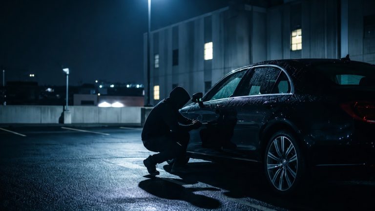 Dark parking garage scene featuring a hooded figure kneeling beside a black car to break in