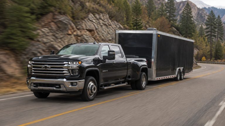 A modern black heavy-duty pickup truck drives along a mountain highway, towing a large cargo trailer