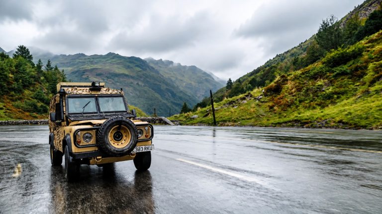 Tan military style off-road vehicle driving on a wet mountain road under cloudy skies