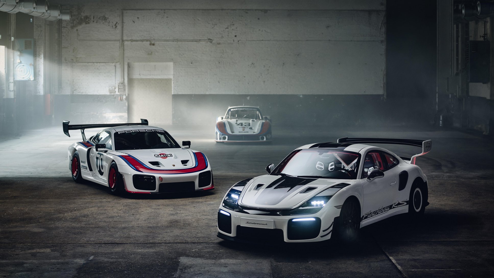 Two white Porsche race cars with Martini liveries parked inside a dimly lit industrial warehouse