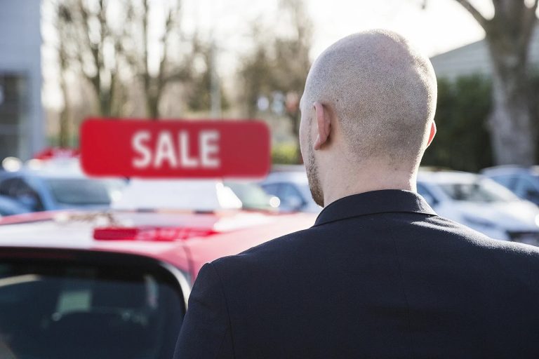 Dark-suited individual viewing a red car with a sale sign at an outdoor dealership lot