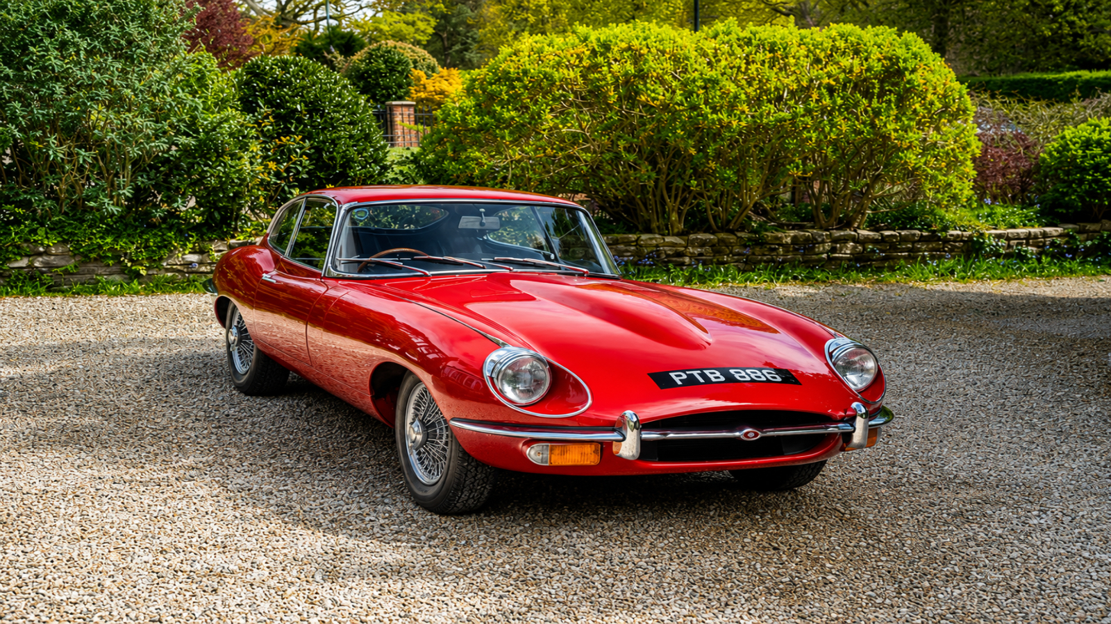 Red Jaguar E-Type classic sports car parked on a gravel driveway with green hedges and gardens in the background.