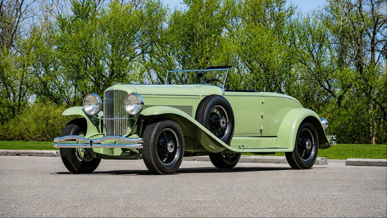 light green vintage Duesenberg is parked on a paved road.