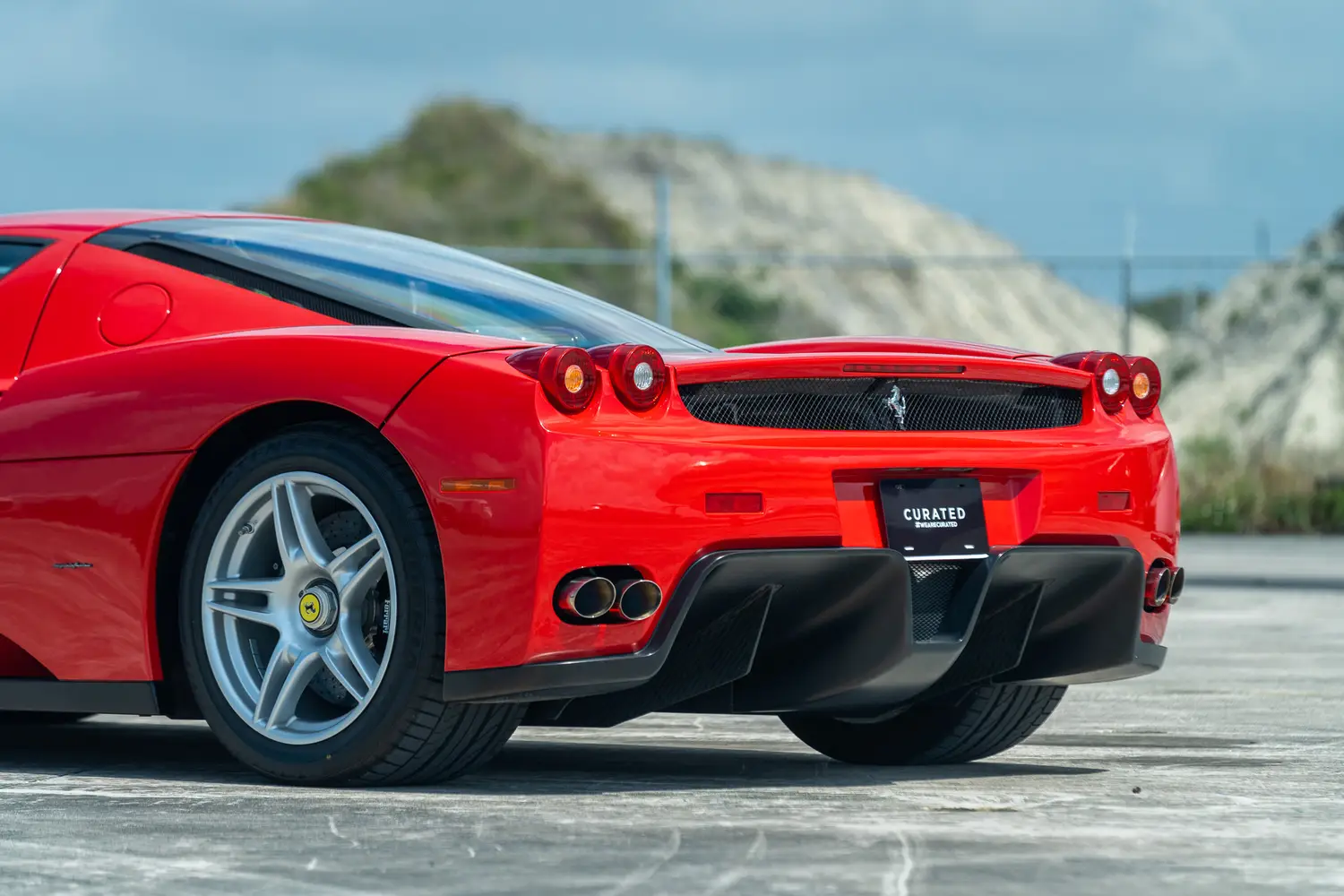 The back of a red Enzo Ferrari is shown on a road in front of a small hill