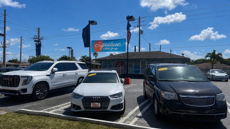 car dealership lot featuring white, black, and other cars parked under a sunny sky