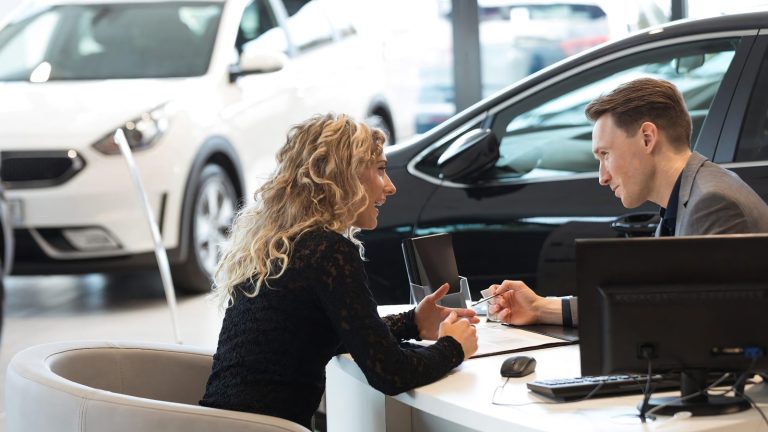 A car salesman and a woman sitting at a desk discussing paperwork in a dealership