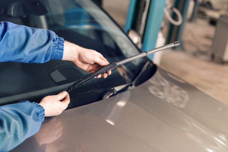 A mechanic in a blue uniform replaces a windshield wiper on a grey car