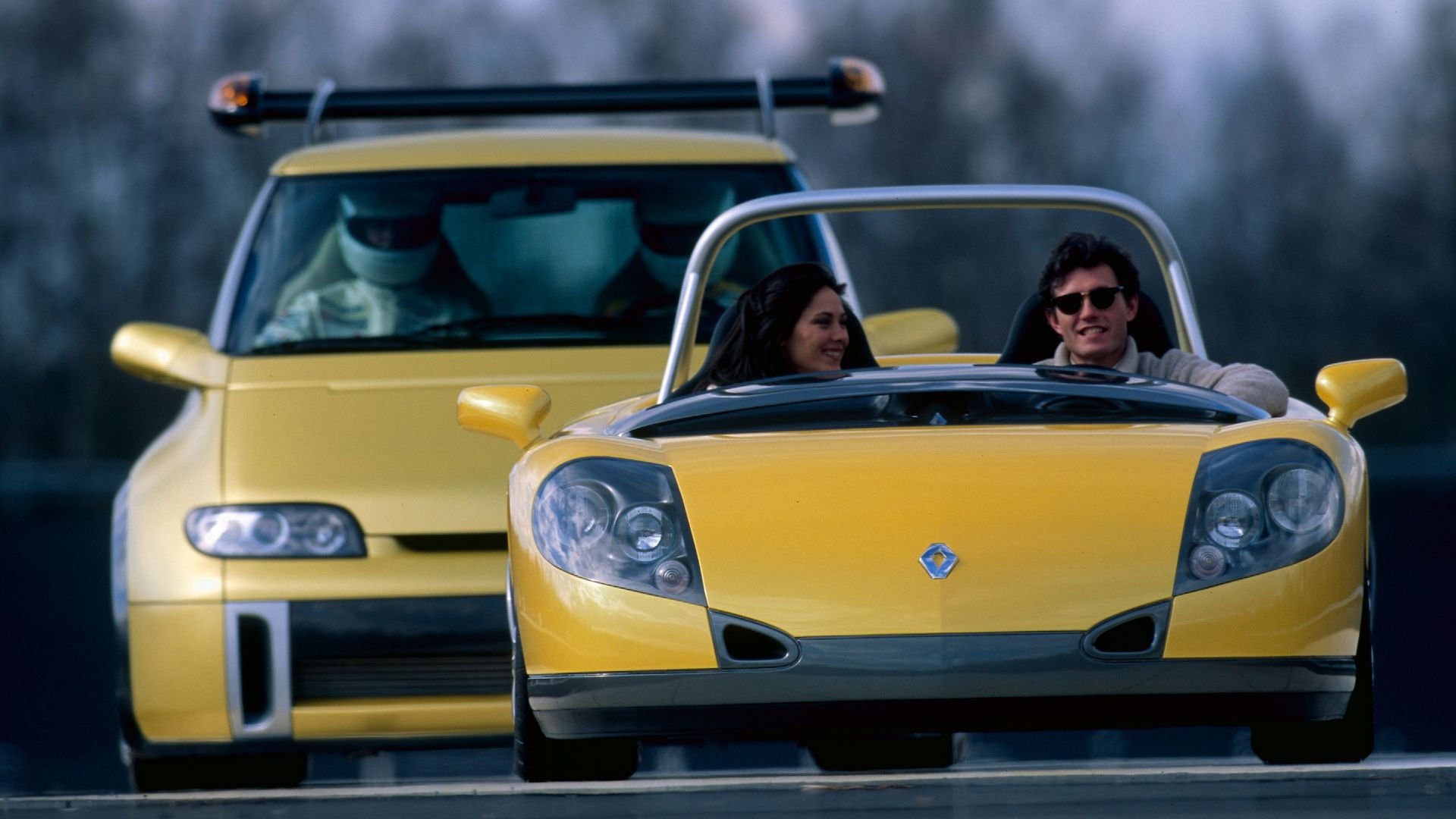 Two people smiling in a bright yellow convertible sports car with another yellow vehicle following behind