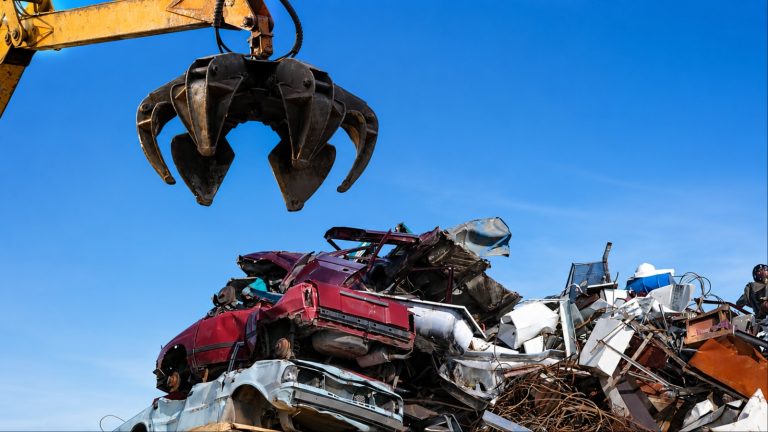 A scrapyard crane claw hangs above a pile of crushed and discarded vehicles