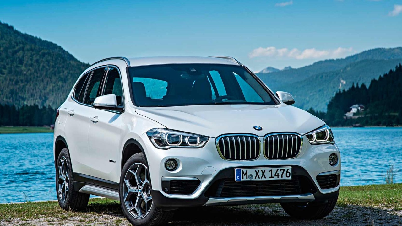A white BMW X1 is parked beside a scenic alpine lake with mountains in the background
