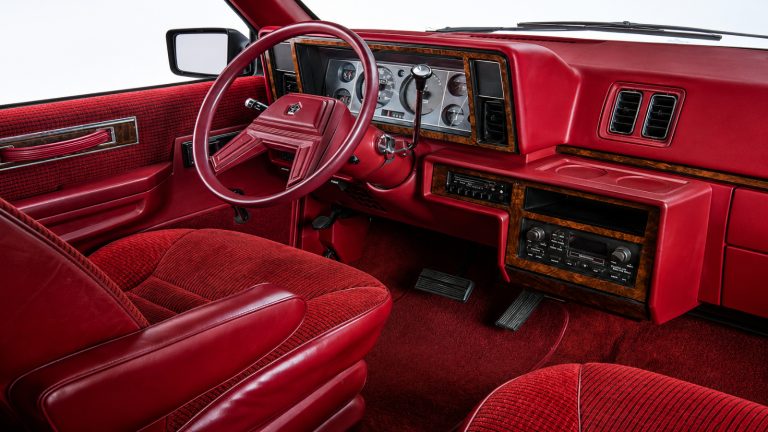Red vintage vehicle interior with a steering wheel, analog gauges, wood-trimmed dashboard, column shifter, and plush seats.