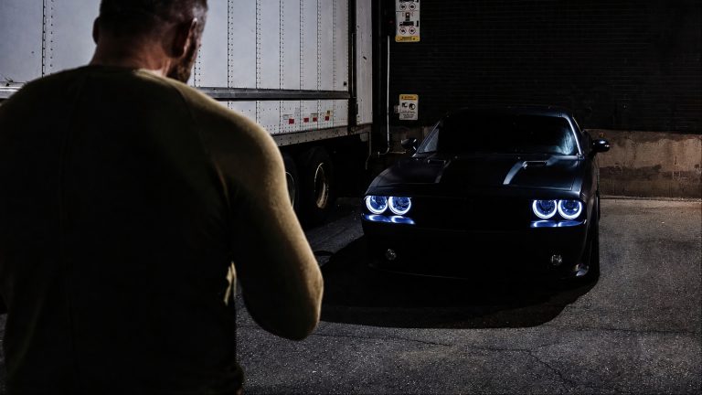 man standing near a semi-trailer looks at a dark Dodge Challenger at night
