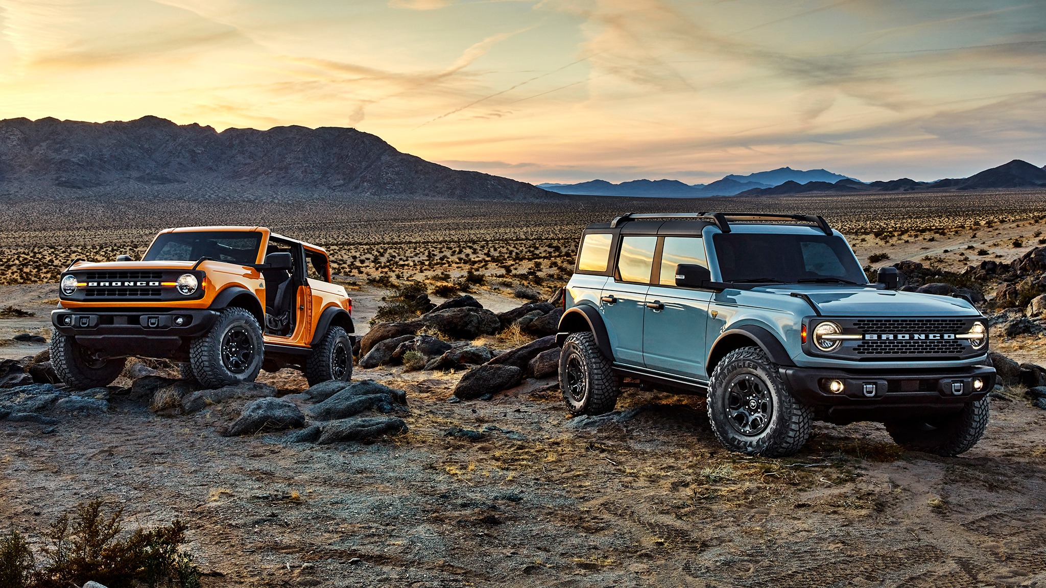 An orange two-door Ford Bronco and a light blue four-door Ford Bronco parked on rocky terrain in a desert landscape at sunset, with mountains in the background.