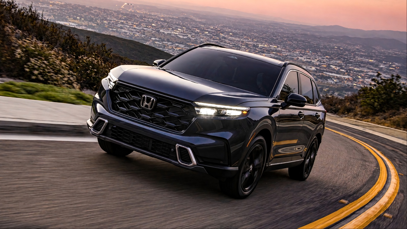 Honda CR-V driving along a winding mountain road at sunset, with city lights below
