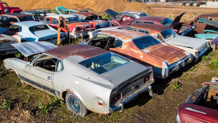 Rusty classic cars sit abandoned in a crowded salvage yard