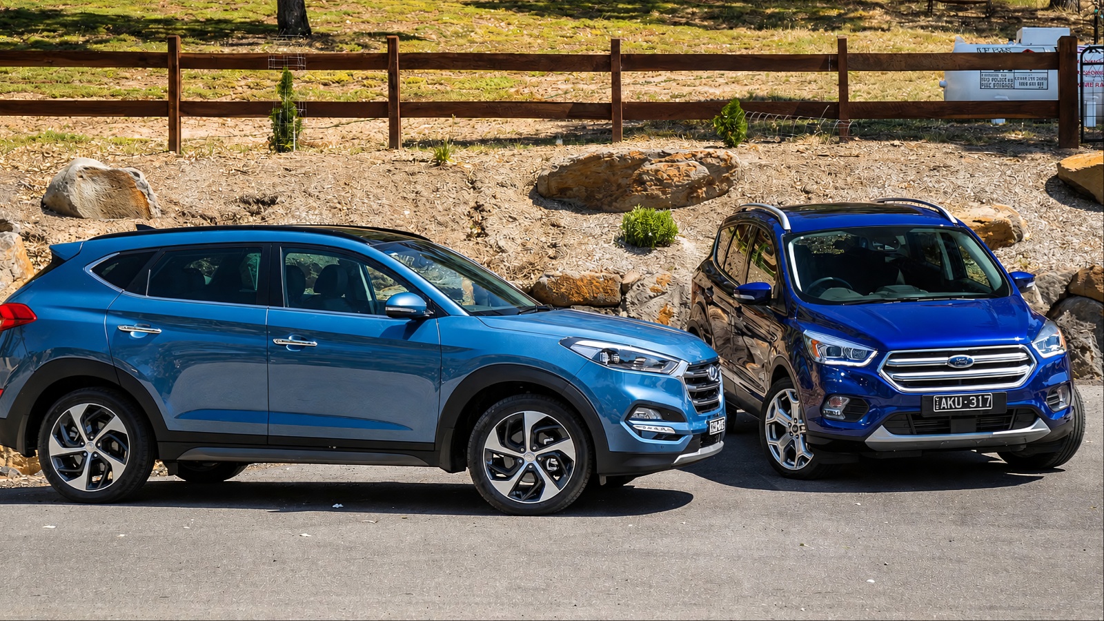 Two blue SUVs parked side by side outdoors near a fence