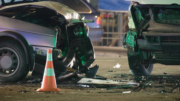 Two heavily damaged vehicles sit at a nighttime crash scene, with debris scattered around a traffic cone