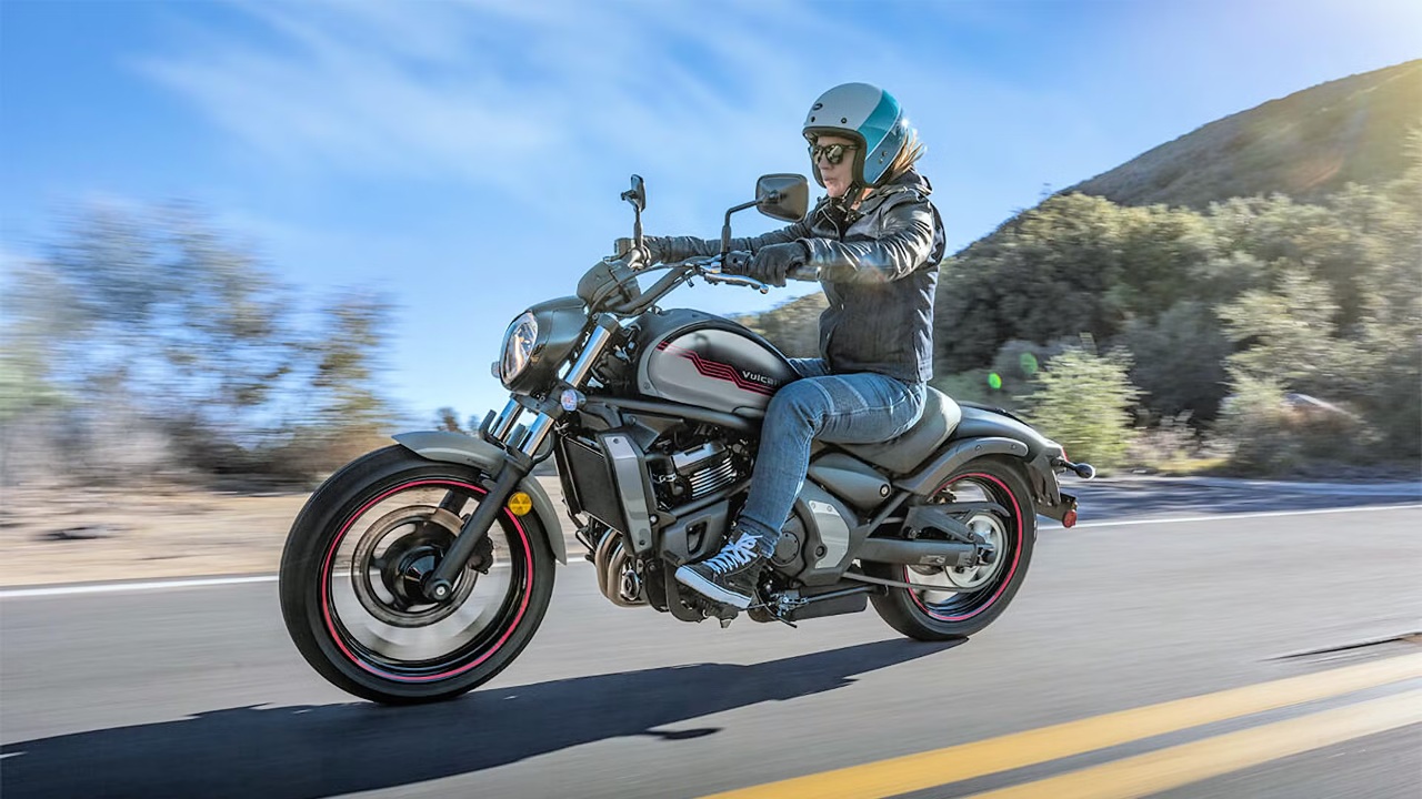 A rider wearing a helmet and protective gear riding a Kawasaki Vulcan S on a scenic mountain road
