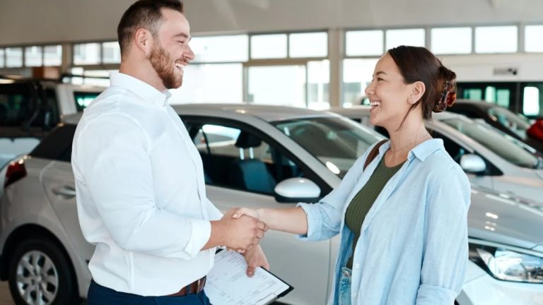 Car dealership salesperson and customer shaking hands beside a newly purchased vehicle
