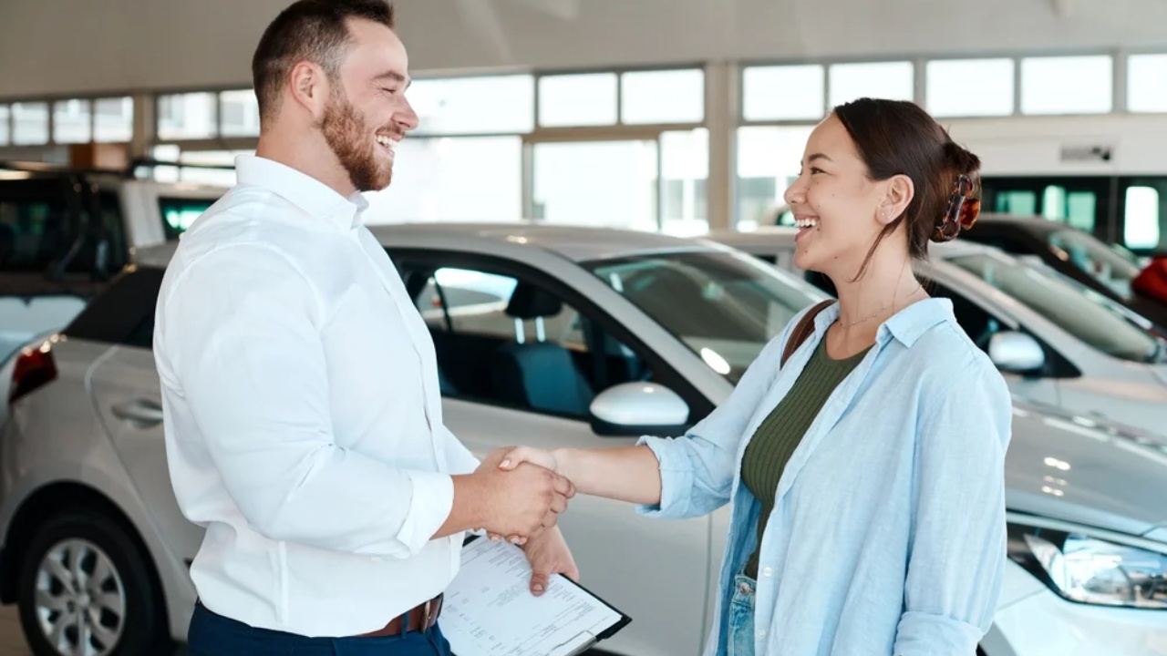 Car dealership salesperson and customer shaking hands beside a newly purchased vehicle