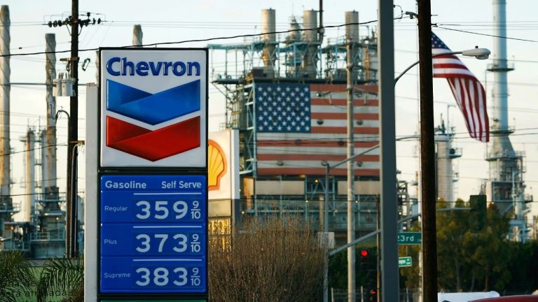 Chevron gas station sign displaying fuel prices with an American flag and refinery in background