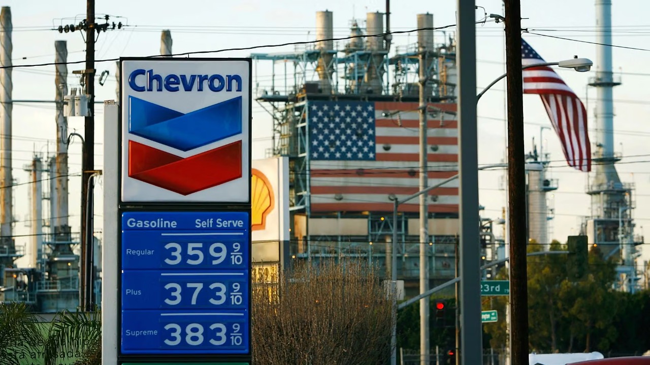 Chevron gas station sign displaying fuel prices with an American flag and refinery in background