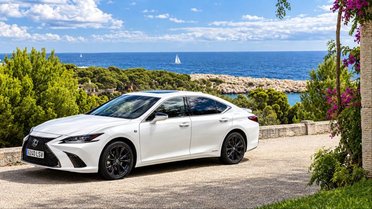 A spotless white Lexus ES 300h is standing on a rough road surrounded by greenery