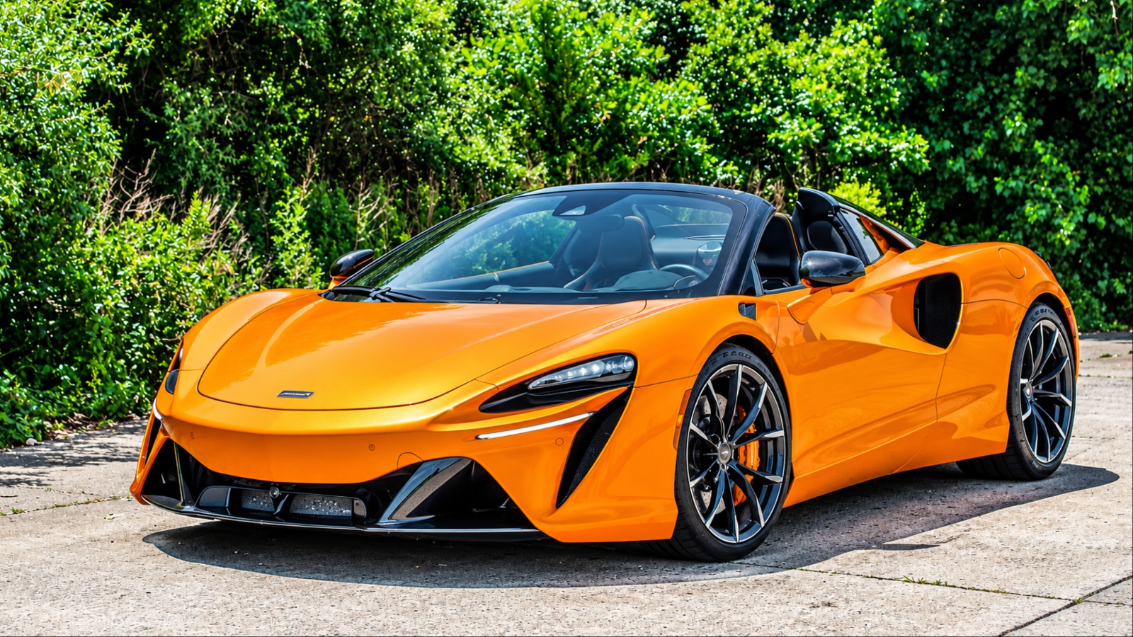 McLaren sports car parked on a sunny road surrounded by lush greenery