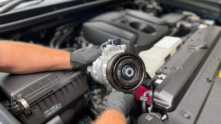 Mechanic holding a replacement AC compressor over a vehicle engine bay