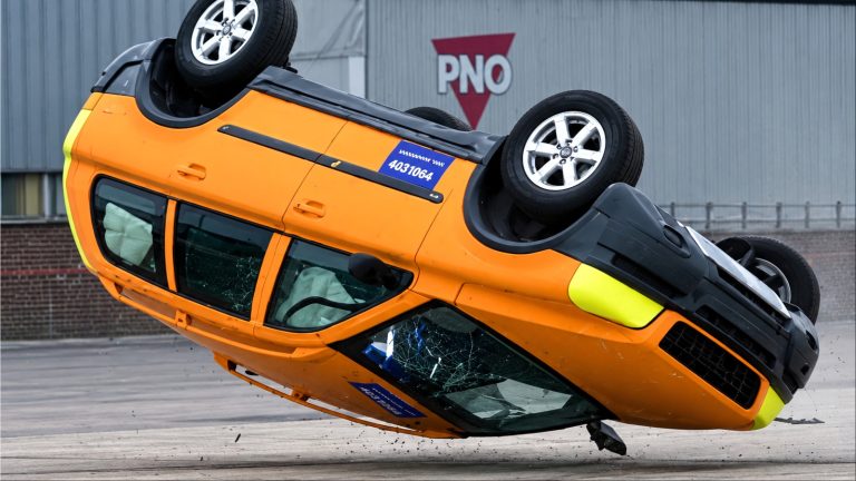Overturned orange van in mid-air crash test with broken windows