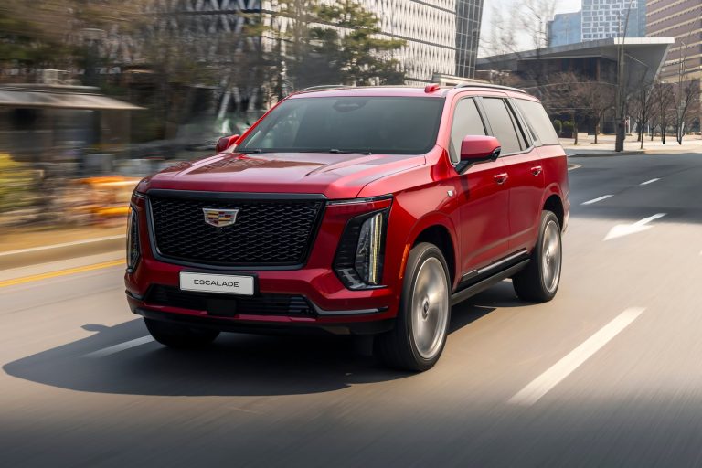 2027 Cadillac Escalade-V driving through a city street, showcasing its bold grille and imposing luxury SUV styling