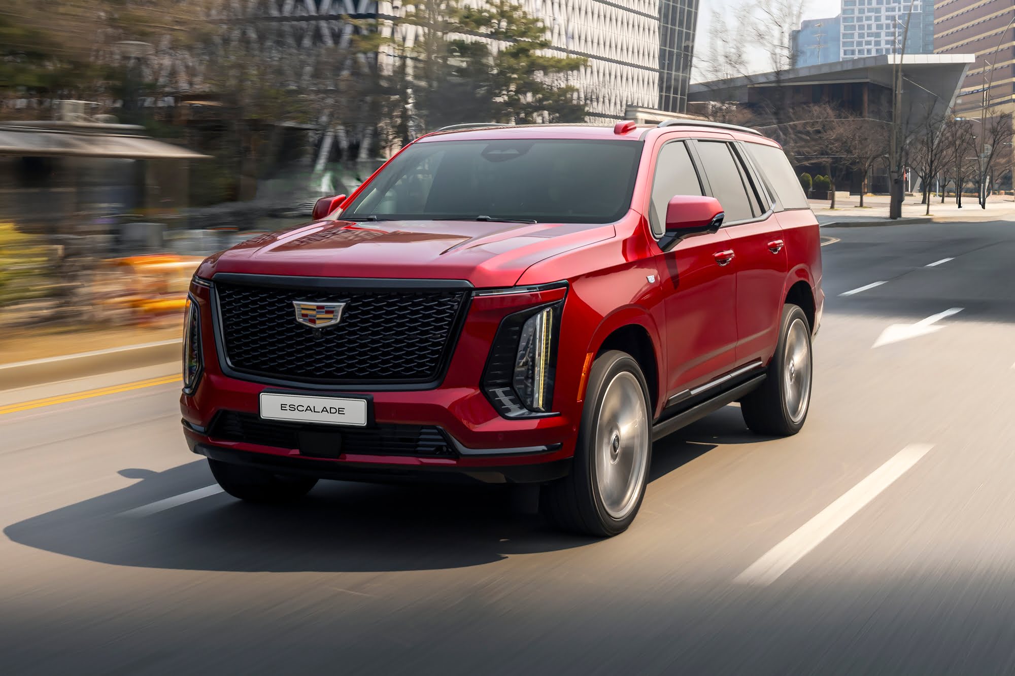 2027 Cadillac Escalade-V driving through a city street, showcasing its bold grille and imposing luxury SUV styling