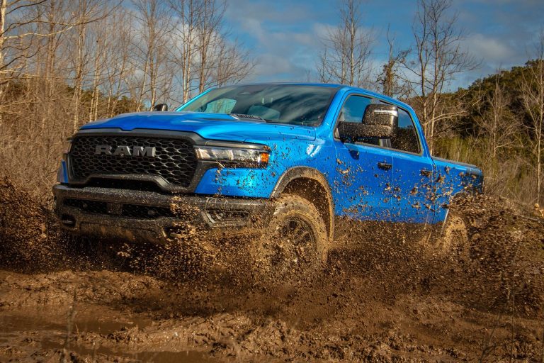 A Ram pickup powers through muddy terrain, spraying dirt across a rugged woodland trail