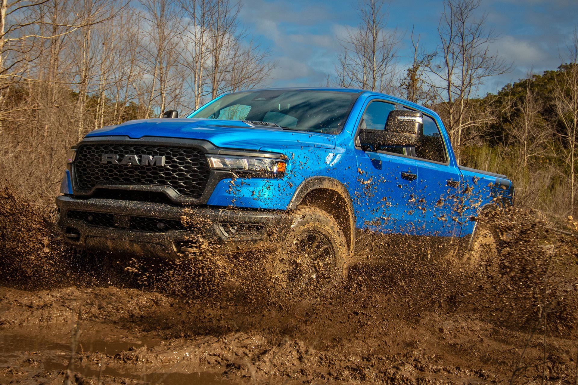 A Ram pickup powers through muddy terrain, spraying dirt across a rugged woodland trail