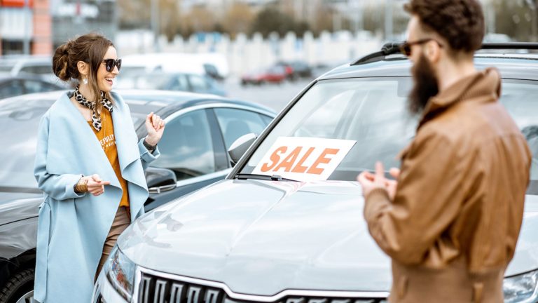 A smiling customer discusses a used car purchase with a seller beside a vehicle marked for sale