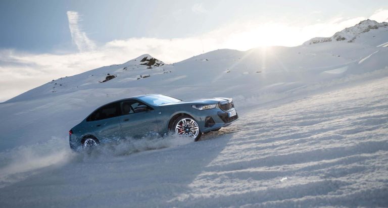 BMW i5 climbs a snowy mountain slope, spraying powder beneath dramatic alpine sunlight