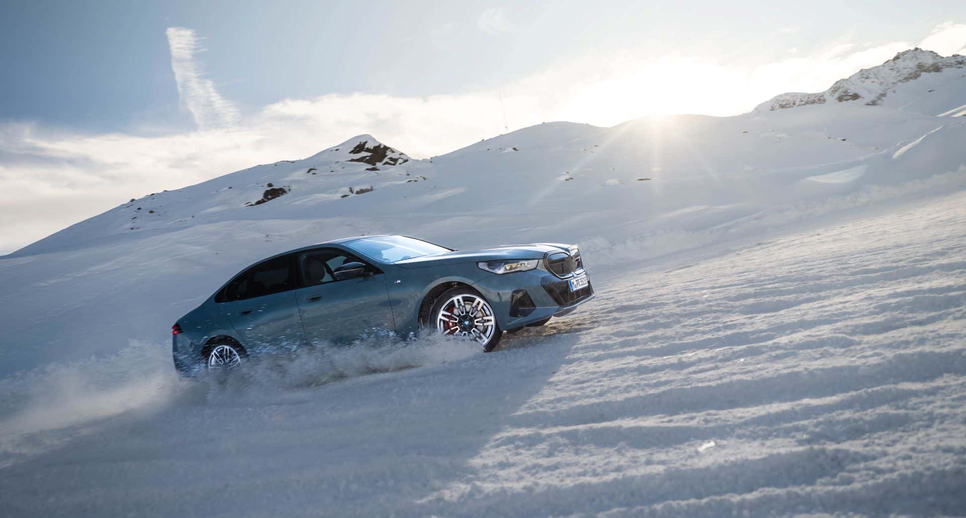 BMW i5 climbs a snowy mountain slope, spraying powder beneath dramatic alpine sunlight