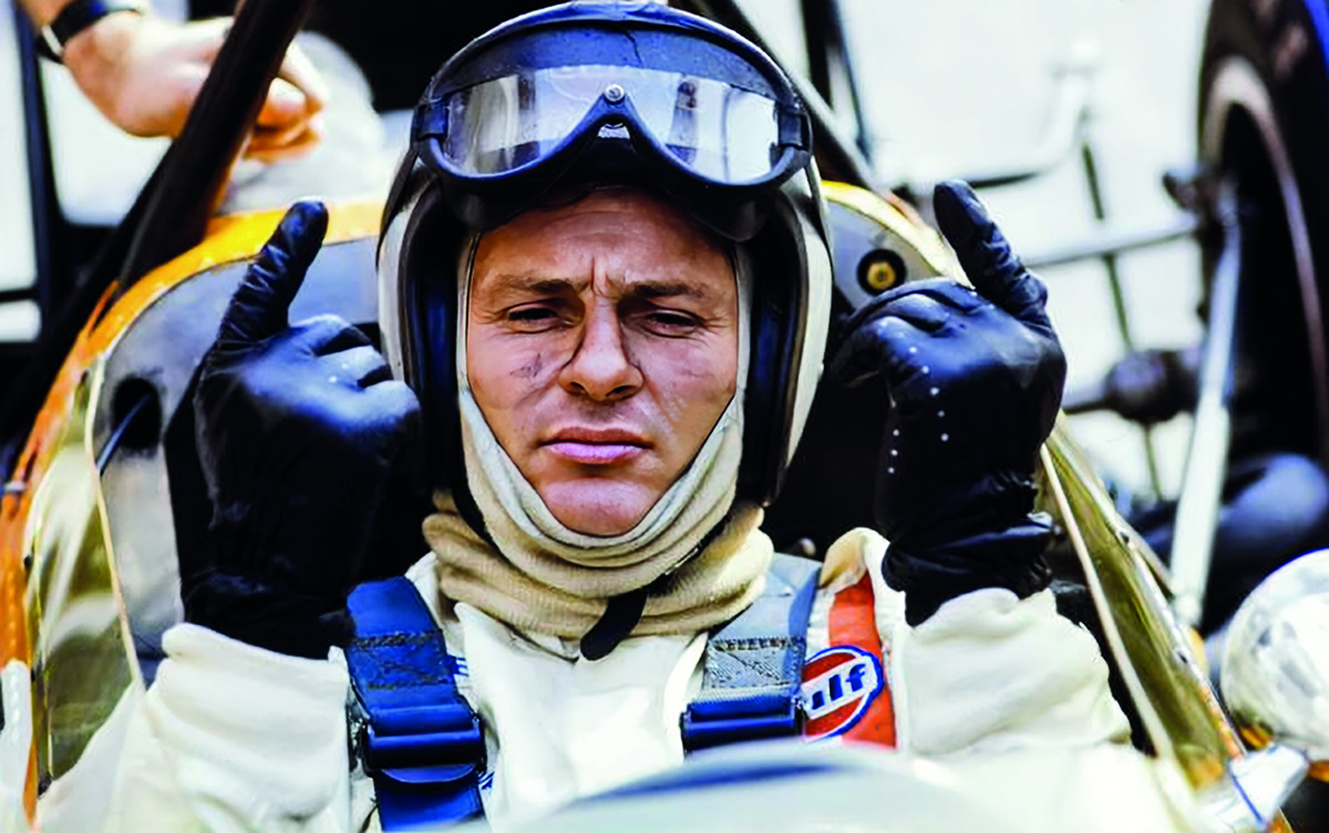 Bruce McLaren in racing gear gives a thumbs-up while seated inside a vintage race car