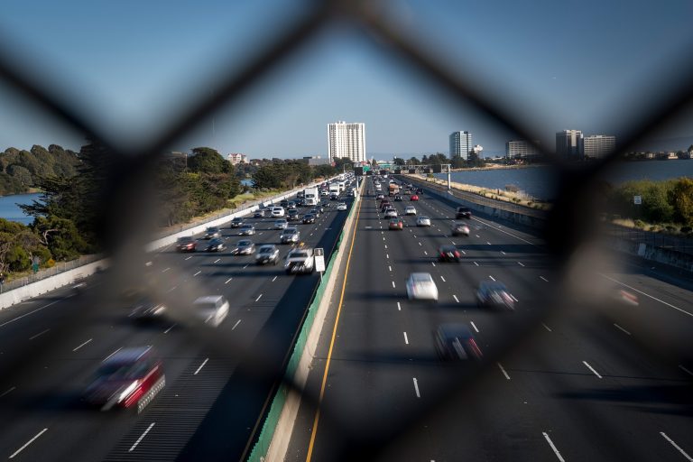 Busy multi-lane highway traffic viewed through a fence, with waterfront scenery and city buildings