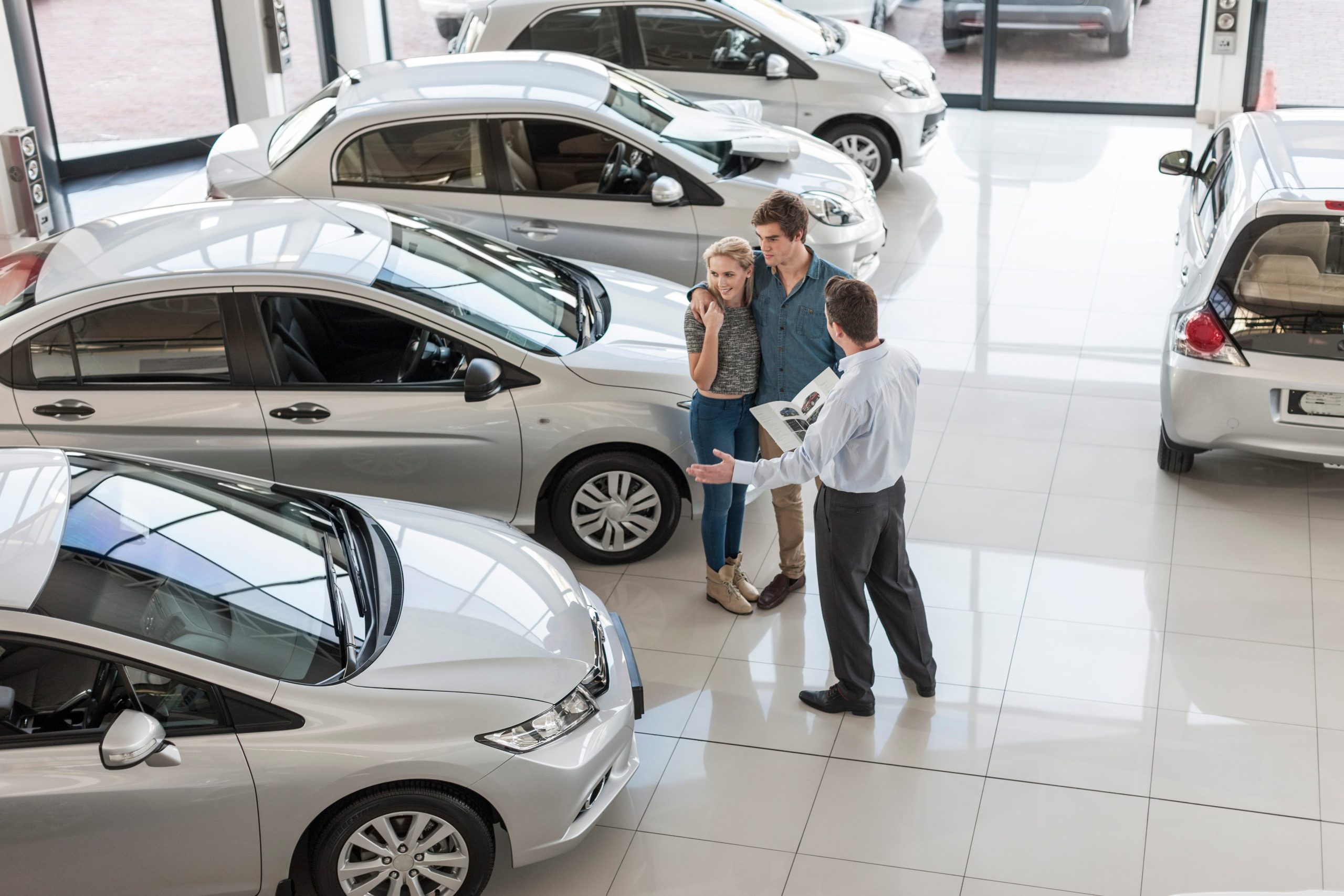 Car dealership salesperson explaining vehicle options to a young couple inside a bright showroom