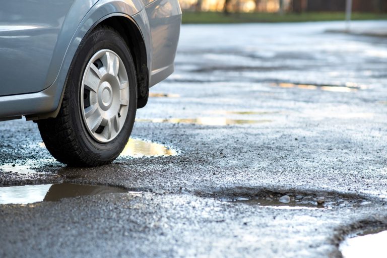 Close-up of a car wheel beside a pothole-filled, waterlogged road, illustrating potential impact damage
